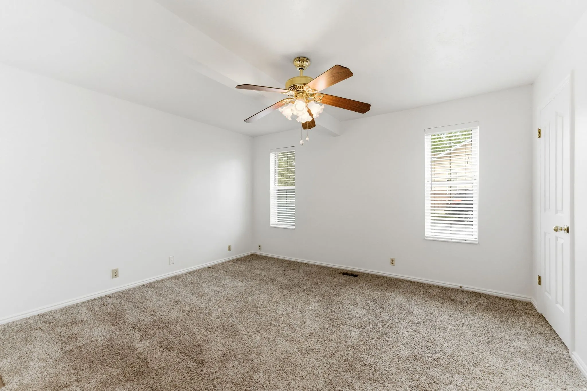 Spacious carpeted room featuring a ceiling fan with light fixture, two windows with blinds, and white walls