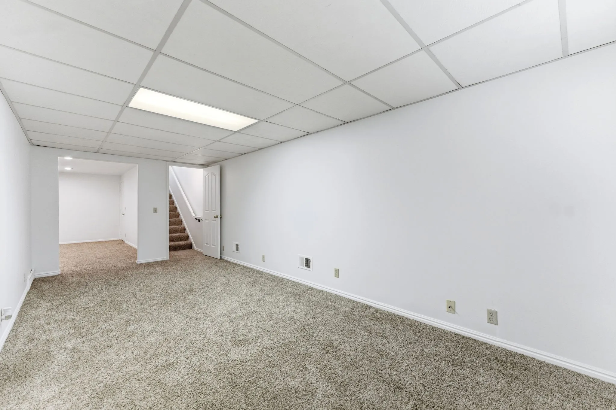 Spacious room featuring neutral carpeting, white walls, a drop ceiling with recessed lighting, and a visible staircase with a white door