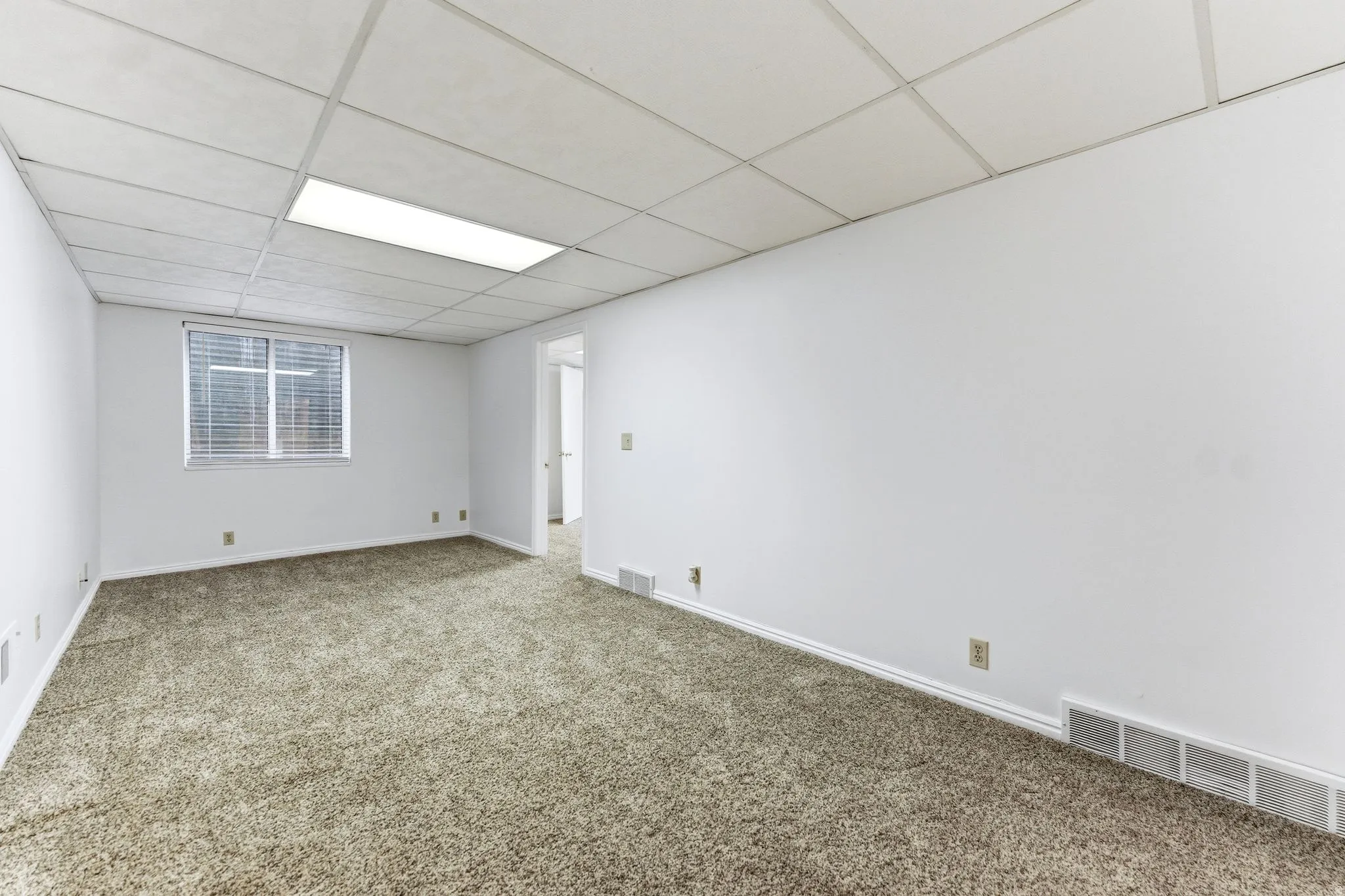 Carpeted room featuring white walls, a grid panel ceiling with recessed lighting, and a window with horizontal blinds