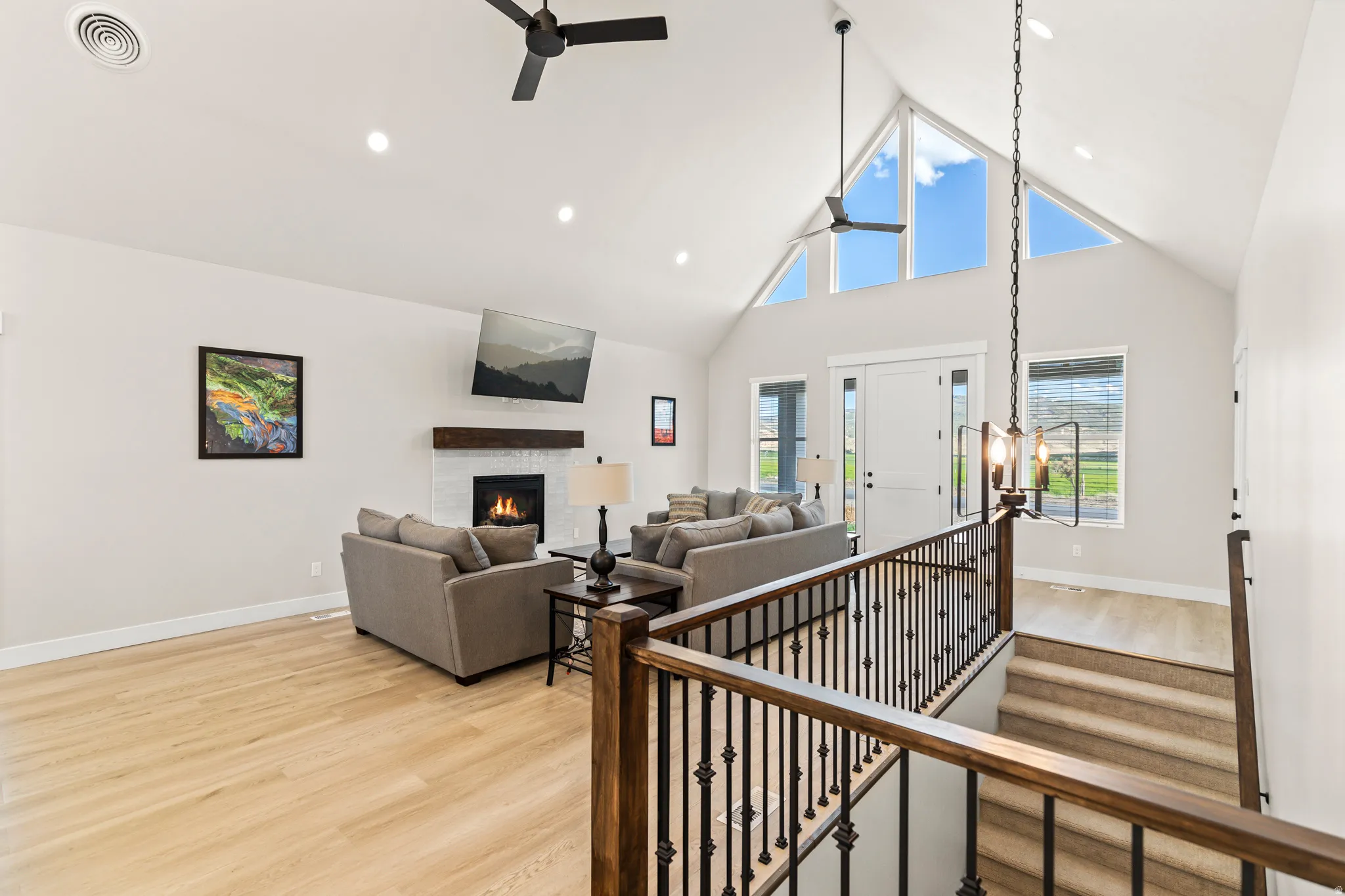 Spacious living area featuring a vaulted ceiling with exposed beams, wood-finish flooring, a white brick fireplace with a wood mantel, and large triangular windows