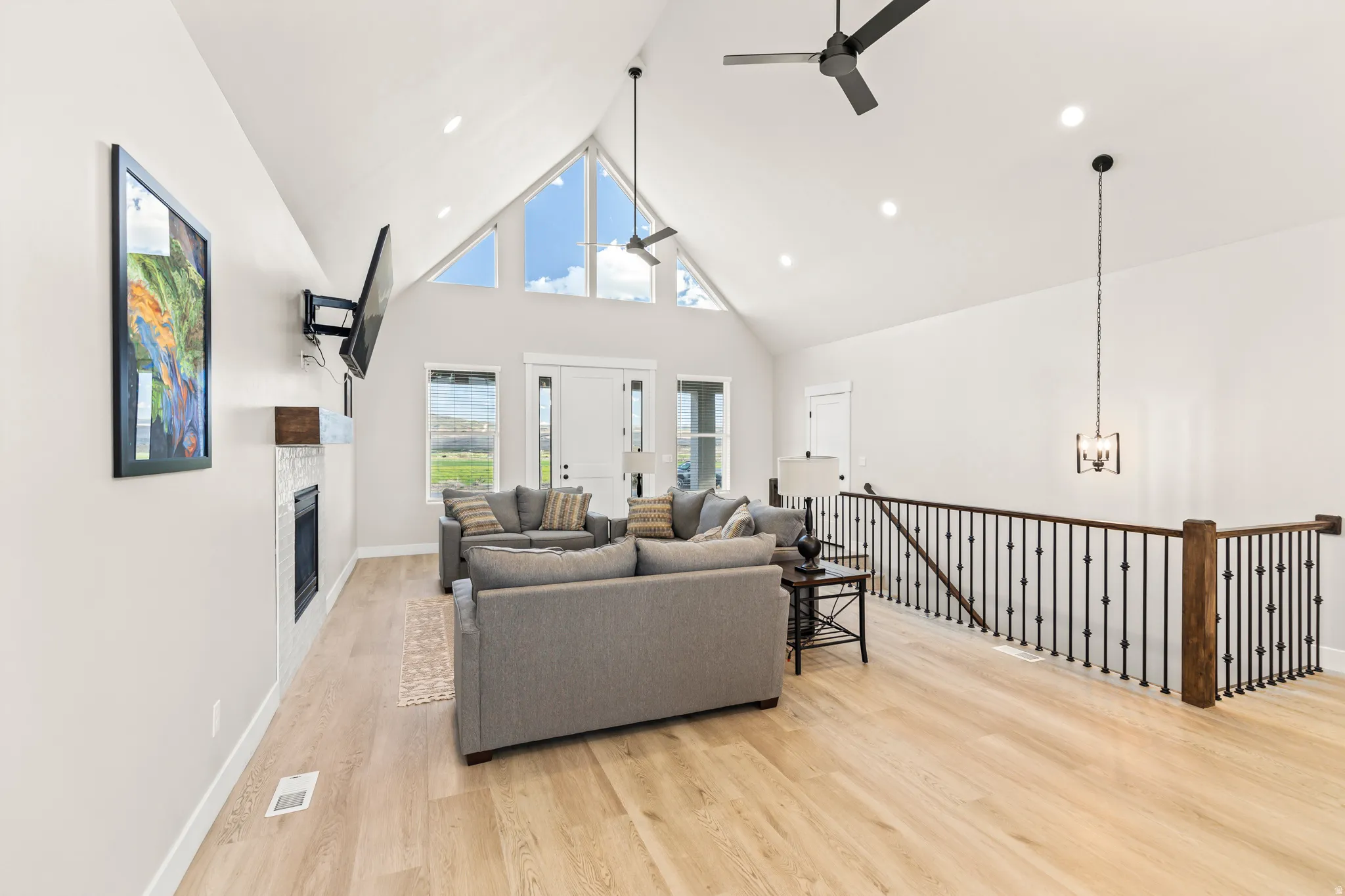 Expansive living area featuring a vaulted ceiling with triangular windows, wood-finish flooring, a contemporary fireplace with a light-colored surround, recessed lighting, and a dark iron spindle railing