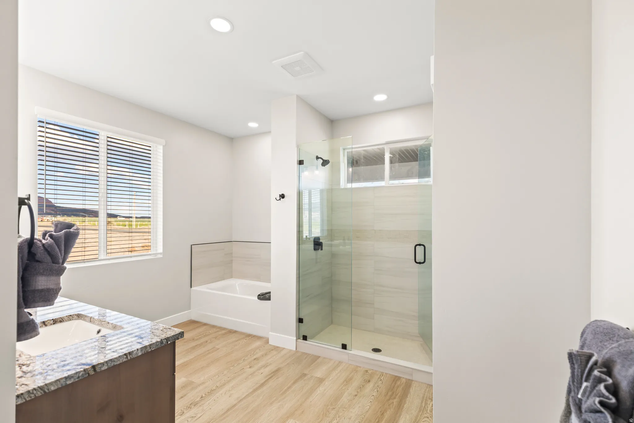 Bathroom featuring a glass-enclosed shower, a built-in tub with tile surround, a vanity with a granite countertop and undermount sink, and wood-finish flooring