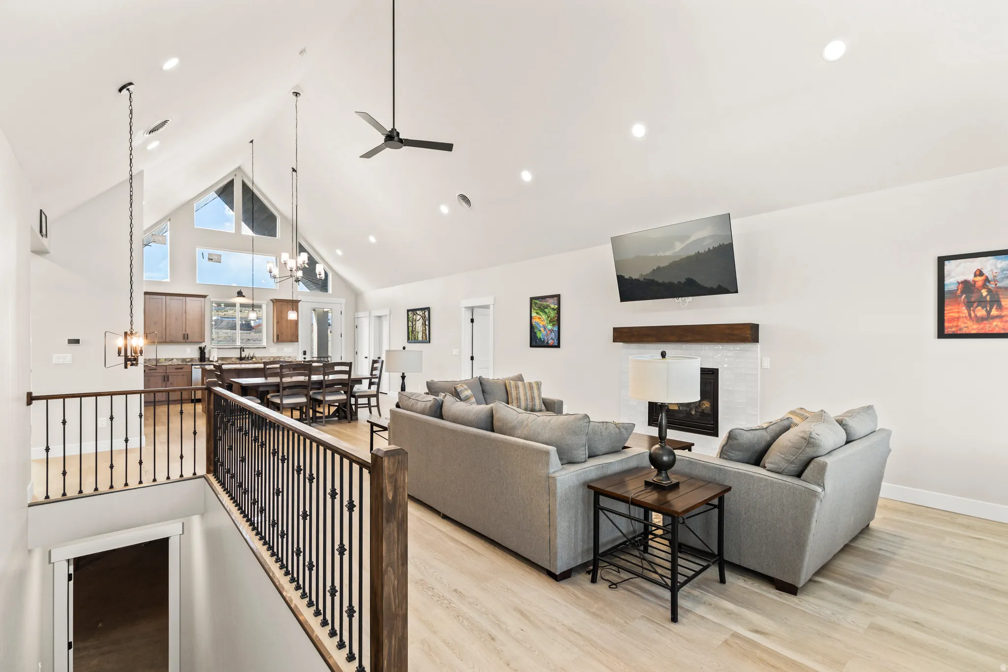Spacious open-concept living area featuring vaulted ceilings, light wood-finish flooring, a white brick fireplace with a wood mantle, recessed lighting, and a kitchen with wood cabinetry and a dining area