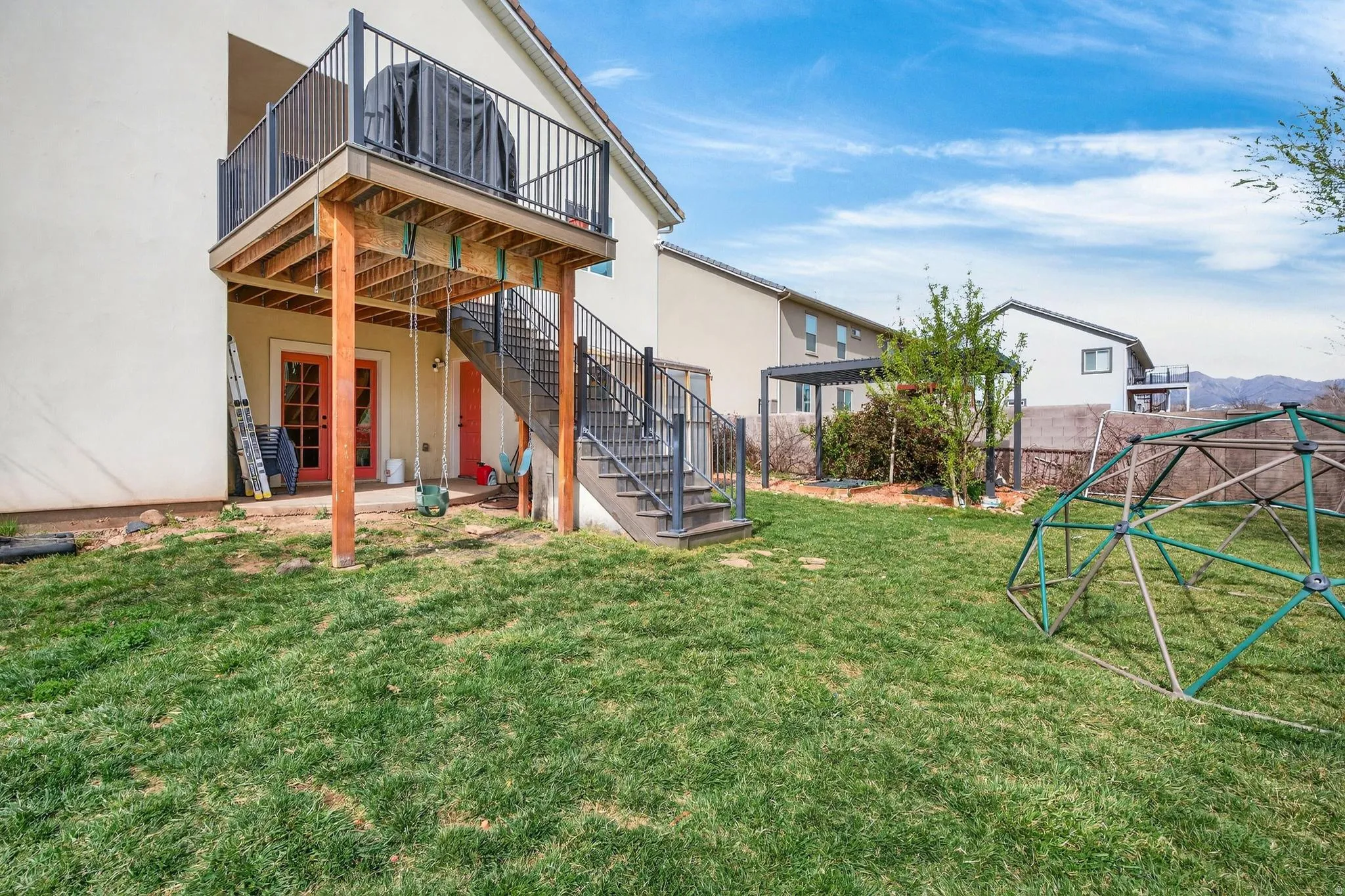 Expansive grass lawn featuring a raised deck with black metal railings, an exterior staircase, and a ground-level patio with double French doors