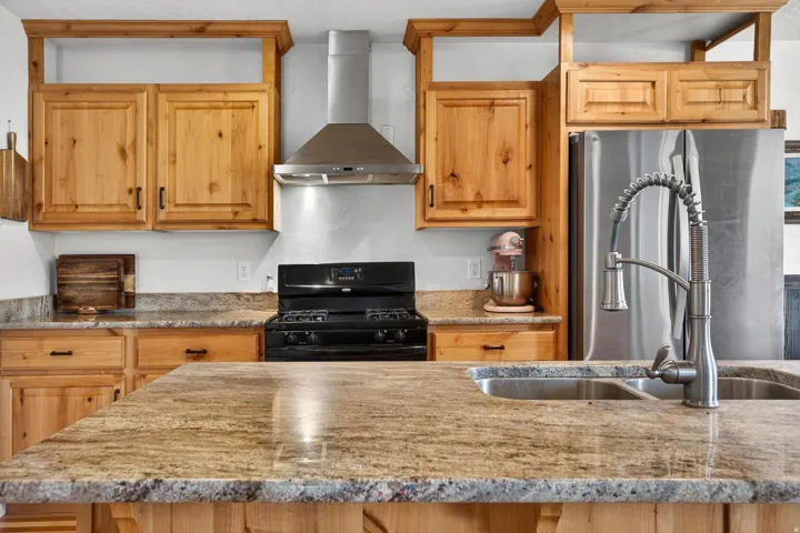 Spacious kitchen featuring natural wood cabinetry, light-toned granite countertops, a stainless steel refrigerator, and a stainless steel gas range with a matching range hood