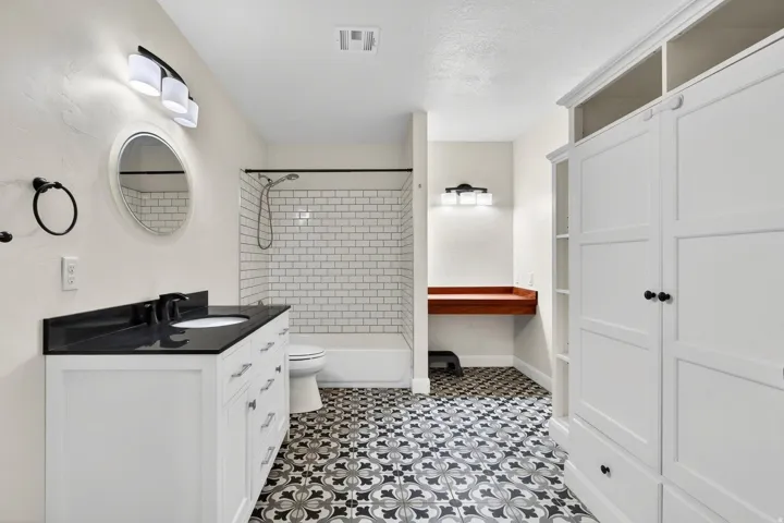 Bathroom featuring patterned floor tiling, a white single vanity with a black countertop, a tub-shower combination with subway tile surround, a built-in storage unit, and a floating wood-finish counter