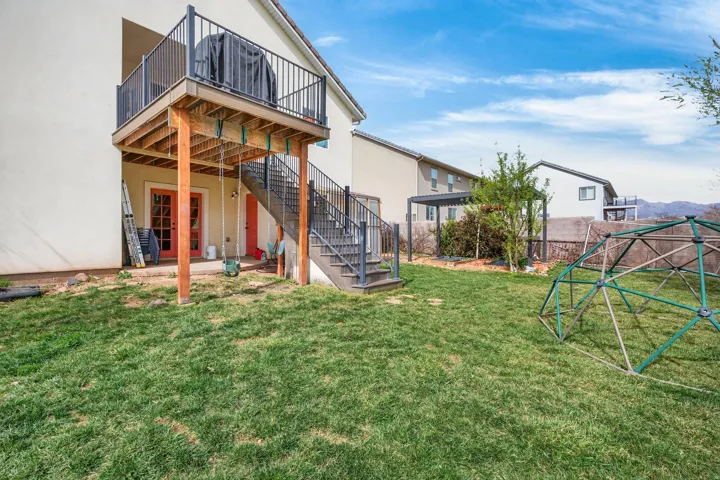 Expansive grass lawn featuring a raised deck with black metal railings, an exterior staircase, and a ground-level patio with double French doors