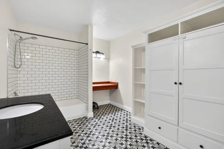Bathroom featuring decorative tiled flooring, a shower-over-tub with subway tile surround, a dark countertop with an undermount sink, a built-in wooden vanity desk, and white cabinetry with open shelving