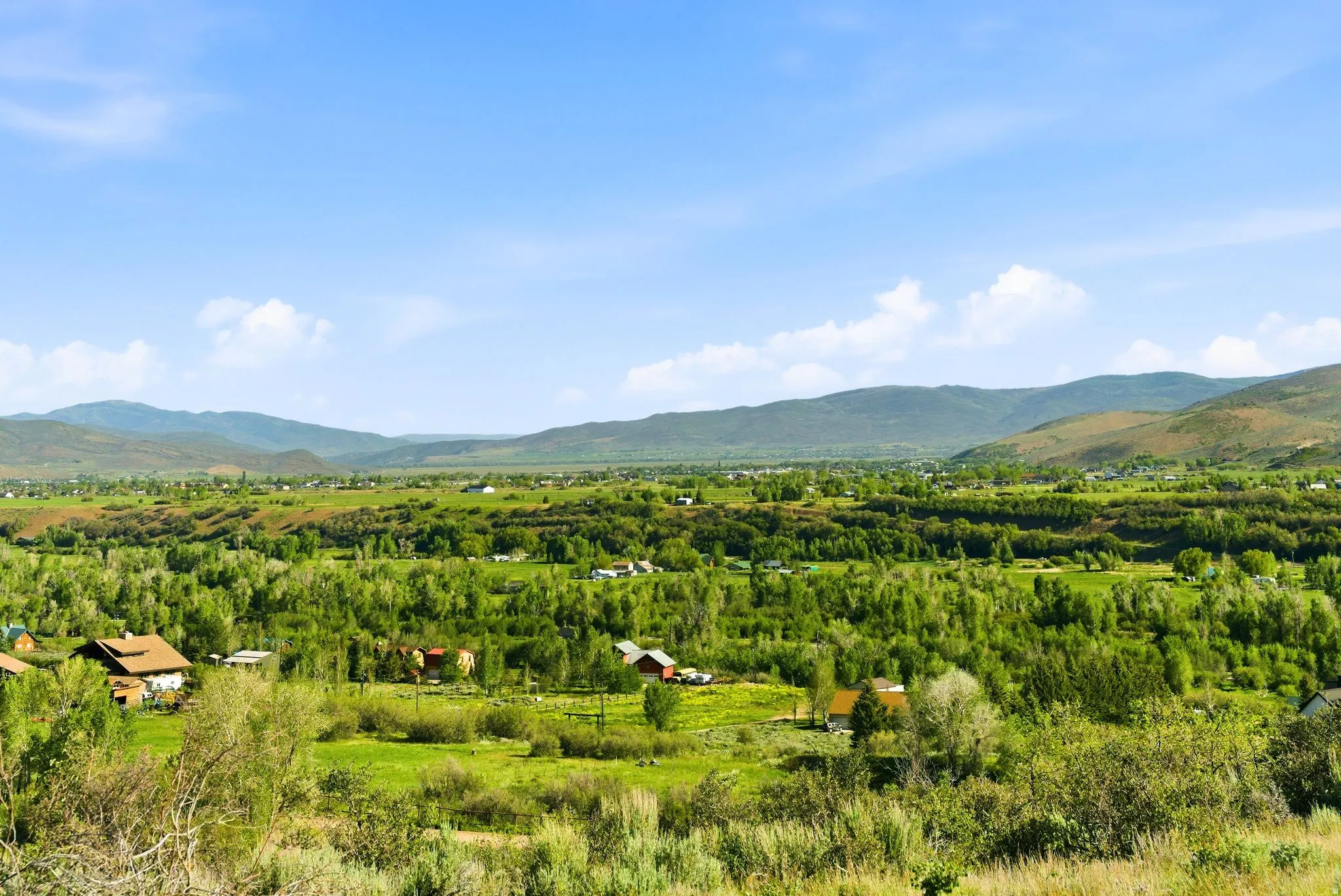 Expansive landscape featuring rolling green hills, a broad valley, and distant mountains under a blue sky