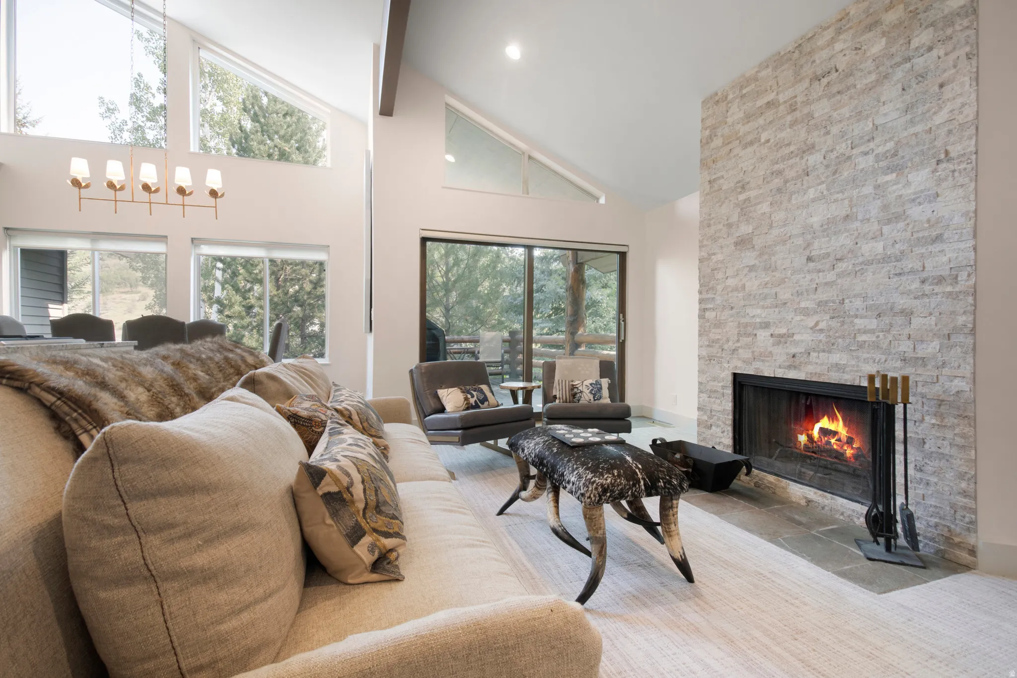 Vaulted ceiling living area featuring a stacked stone fireplace with a black metal firebox, large glass sliding doors, and ample natural light from multiple windows