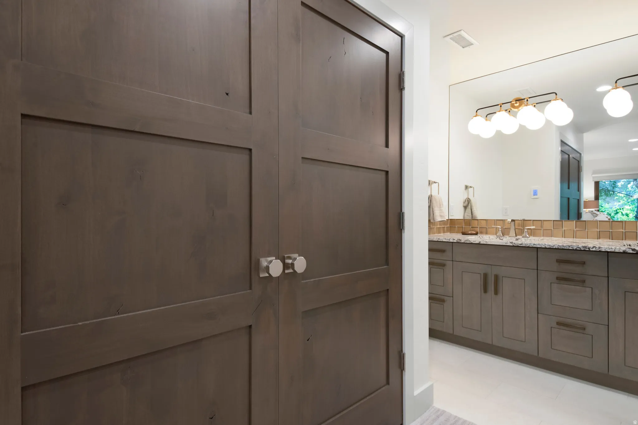 Double bi-fold doors with round brushed nickel hardware, light wood-finish vanity with drawer and cabinet storage, stone countertop, dual undermount sinks, and a large frameless mirror with a multi-bulb vanity light fixture