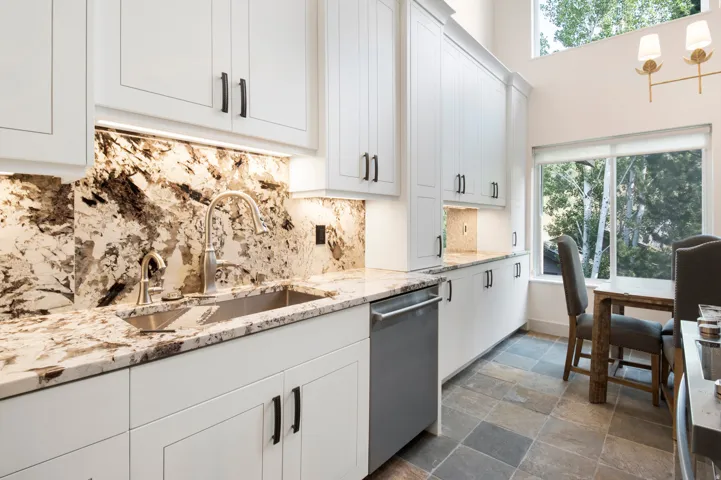 Kitchen featuring white cabinetry with dark hardware, stone countertops and backsplash, an undermount sink with a gooseneck faucet, and slate-style flooring