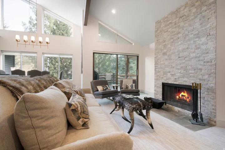 Vaulted ceiling living area featuring a stacked stone fireplace with a black metal firebox, large glass sliding doors, and ample natural light from multiple windows