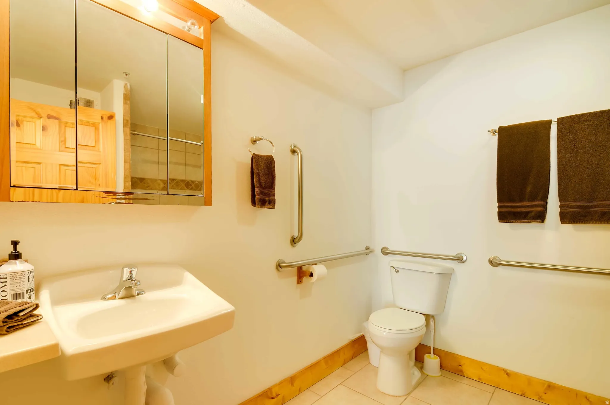 Bathroom featuring a wall-mounted sink, a wood-finish medicine cabinet with mirrored doors, a standard toilet, and neutral-toned floor tiling