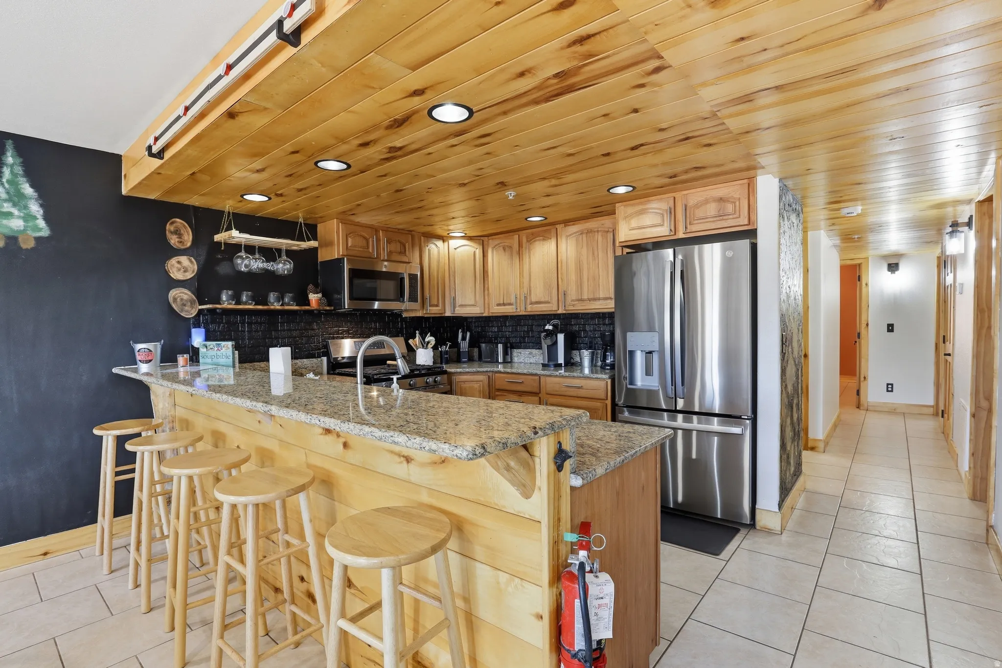 Kitchen featuring a wood-panel ceiling with recessed lighting, wood cabinetry, stainless steel appliances, a breakfast bar with granite-style countertops, and a black tile backsplash