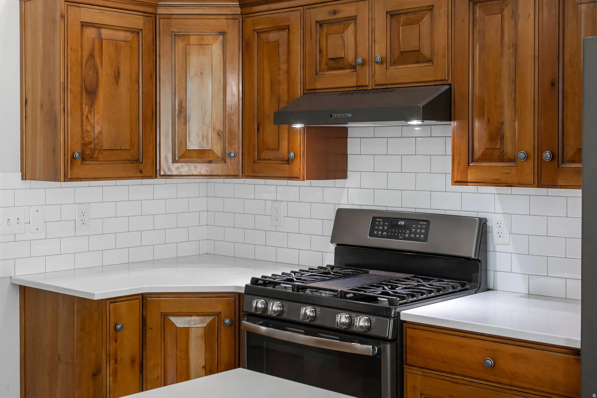 Kitchen featuring wood cabinetry, white subway tile backsplash, light-toned countertops, a stainless steel range, and a matching range hood