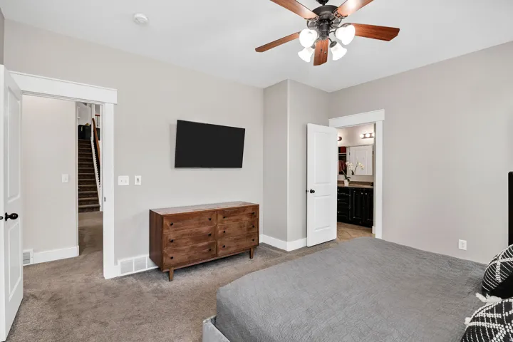 Carpeted room featuring a ceiling fan with integrated lighting, a doorway opening to a staircase, and another doorway revealing a bathroom vanity with a mirror and lighting
