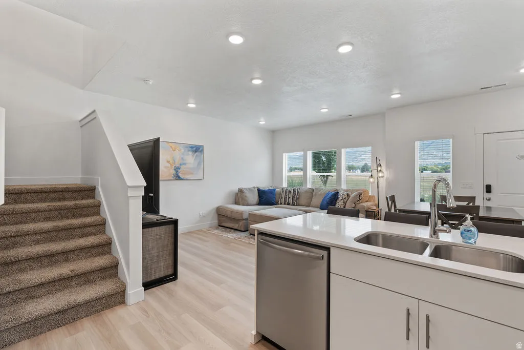 Open-concept living area featuring light wood-finish flooring, recessed lighting, a modern kitchen island with an undermount double sink, and large windows offering exterior views