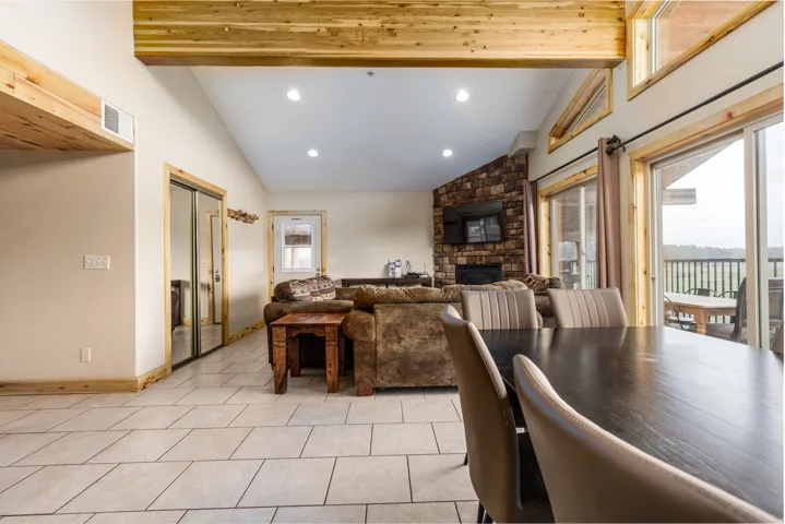 Open-concept living area featuring exposed wood beams, recessed lighting, stone accent wall with fireplace, and a sliding glass door leading to a balcony