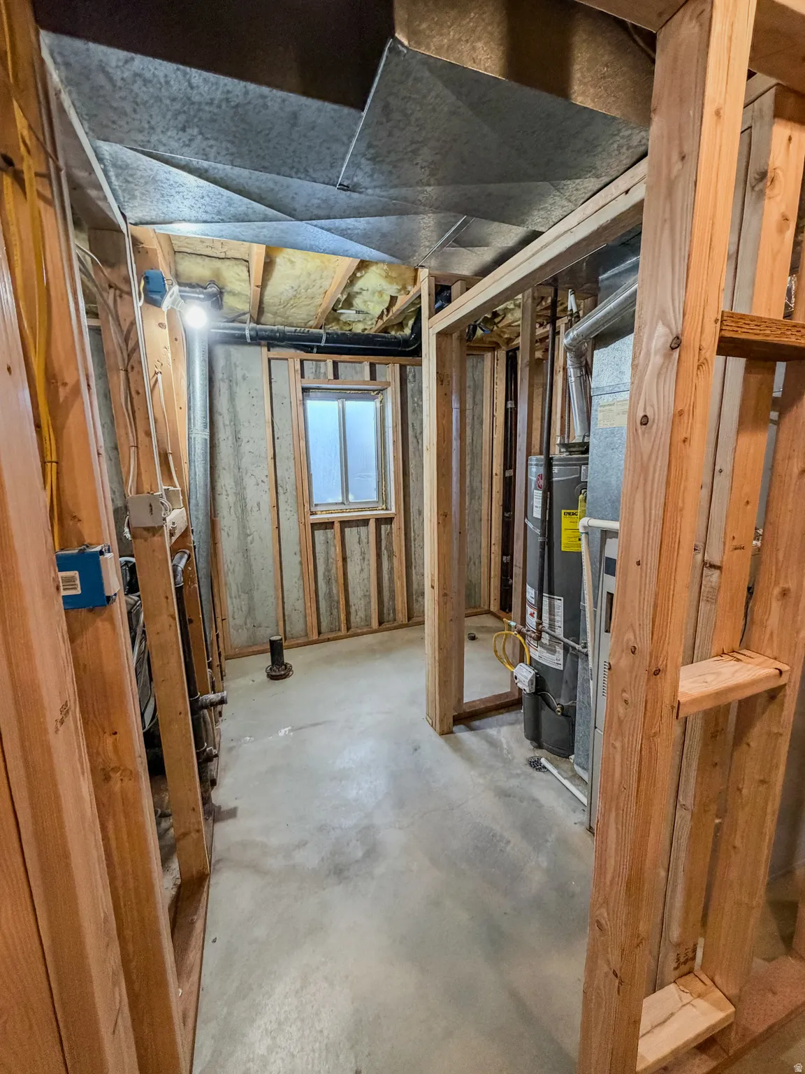 Unfinished basement space featuring exposed wooden framing, concrete flooring, galvanized ductwork, and a small window