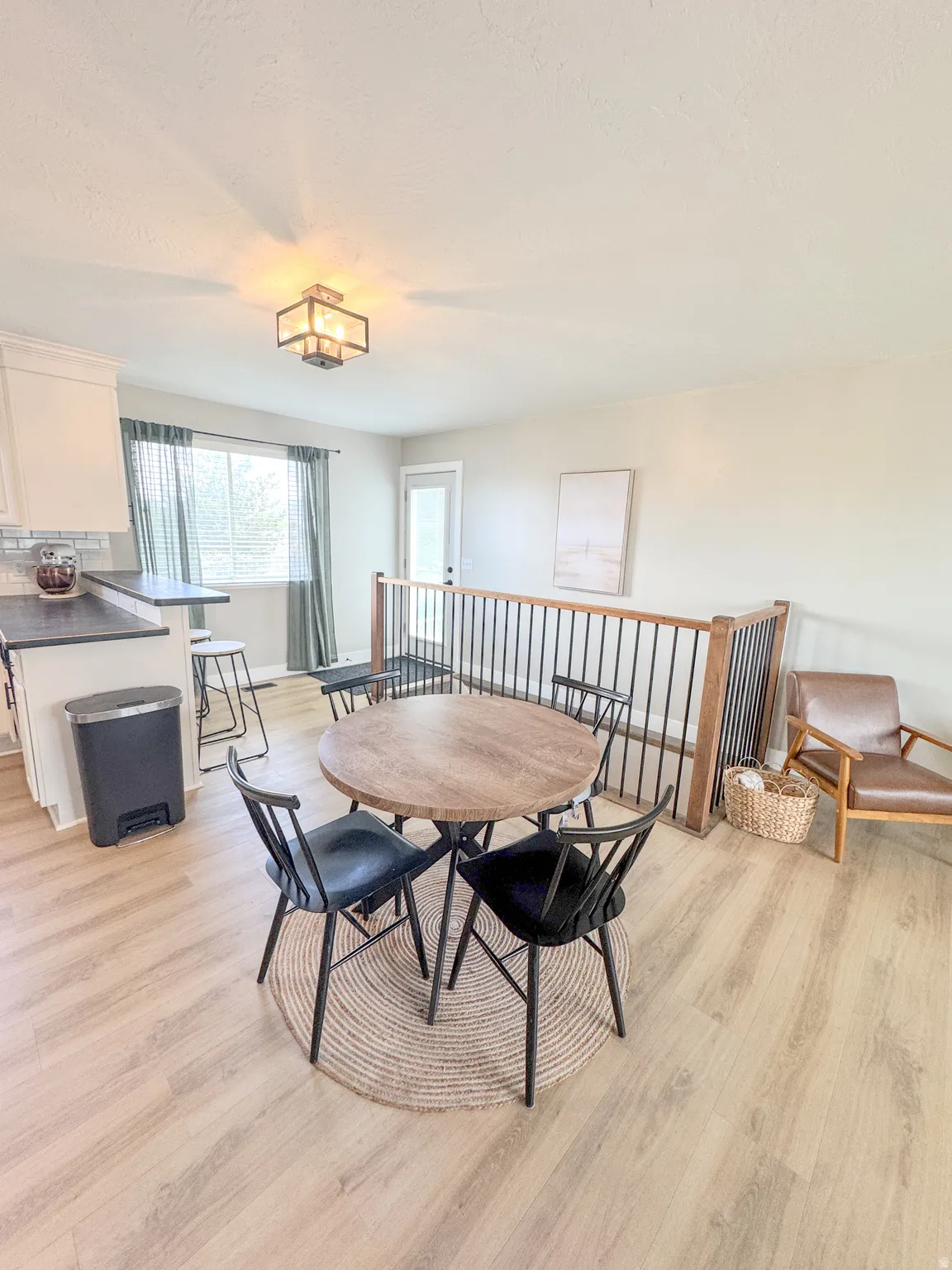 Open-concept living space featuring wood-finish flooring, a contemporary ceiling light fixture, a window with natural light, a white exterior door, and a staircase with a wooden handrail and black metal spindles