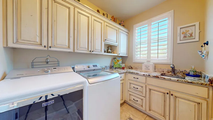 Utility room featuring upper and lower cabinetry with distressed finishes, a built-in sink with a single-handle faucet, a window with plantation shutters, and light-toned flooring