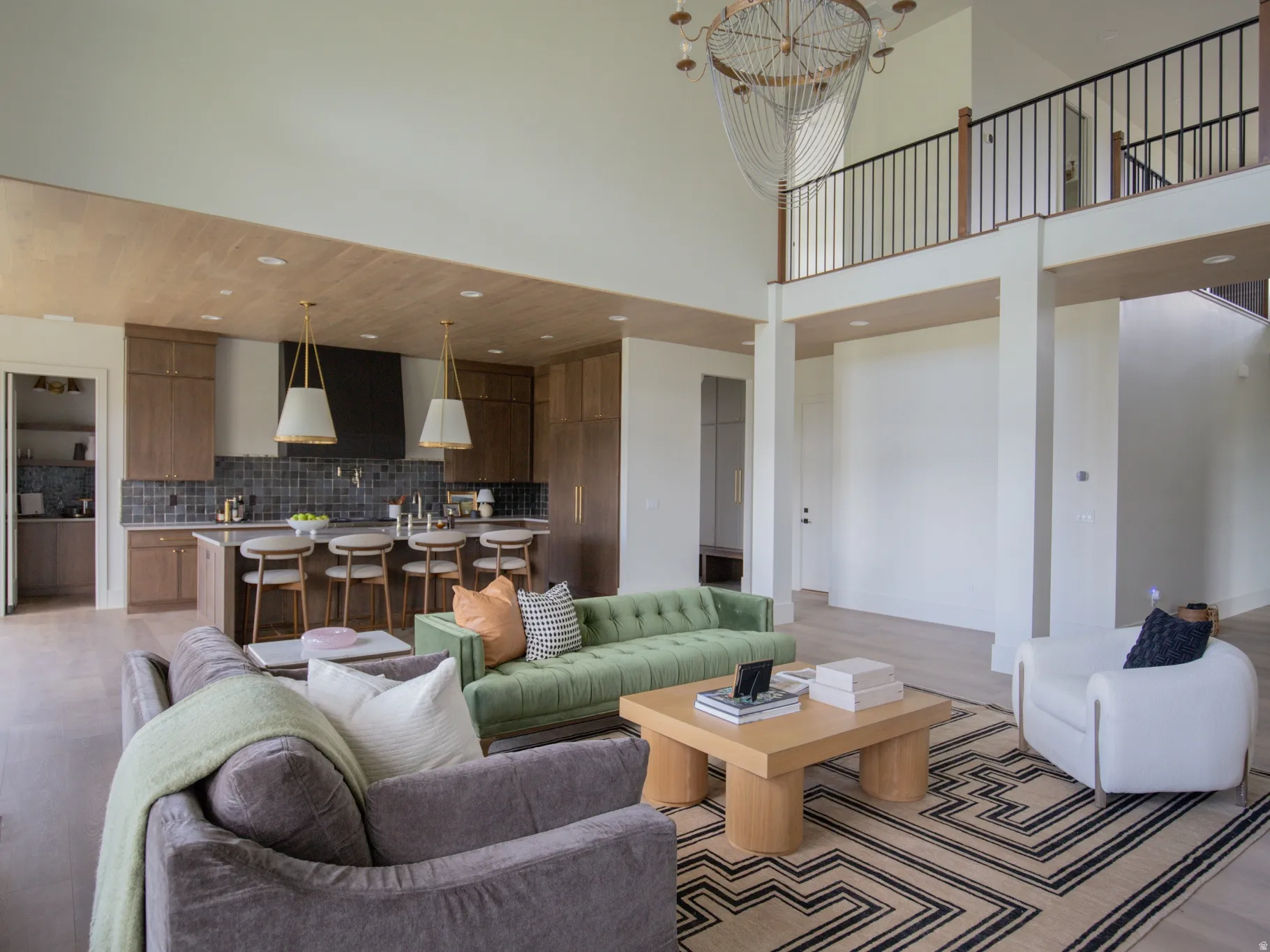 Open-concept living area featuring a double-height ceiling, light wood-finish flooring, a kitchen island with seating, a built-in range hood, and a second-story interior balcony with black metal railings