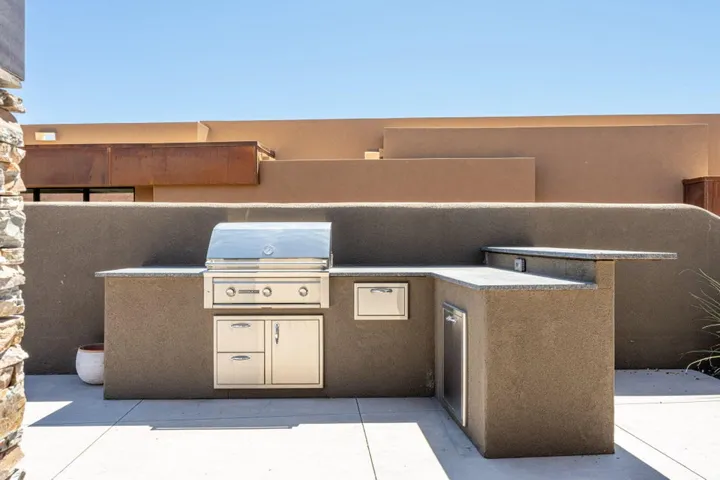 Outdoor L-shaped kitchen featuring a built-in stainless steel grill, integrated storage drawers, a compact refrigerator, and a granite countertop