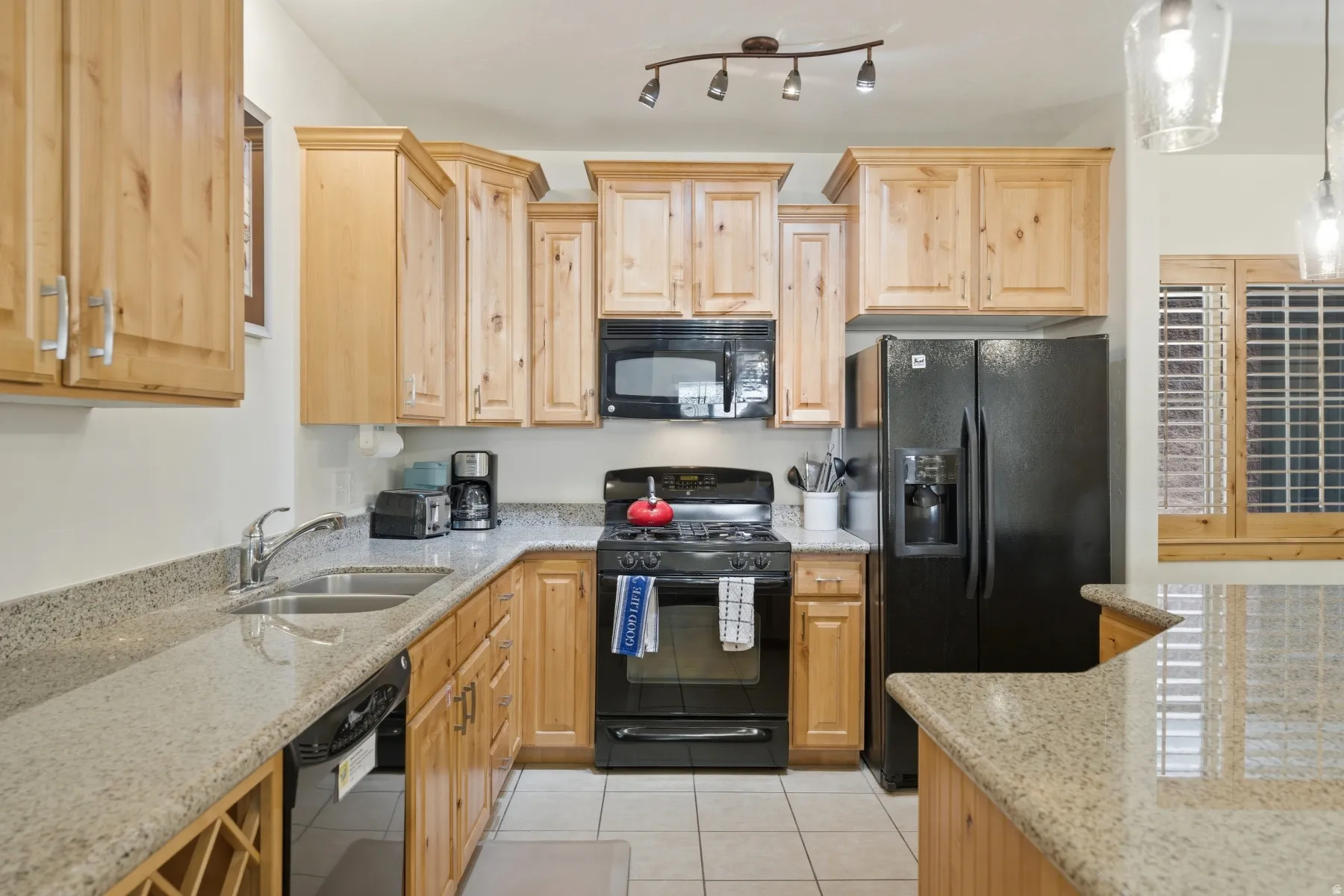 Kitchen featuring light wood cabinetry, stone countertops, black appliances, a double basin sink, and tile flooring