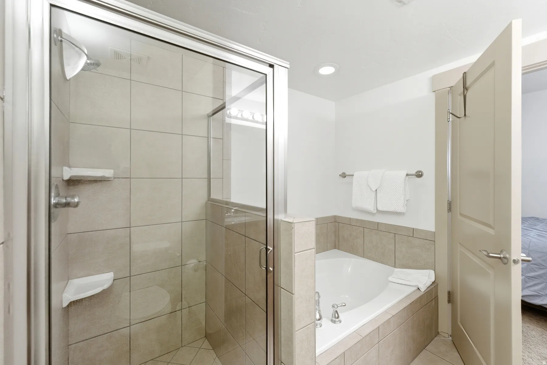 Walk-in shower with clear glass enclosure and light-toned tile surround, accompanied by a built-in corner soaking tub with matching tile