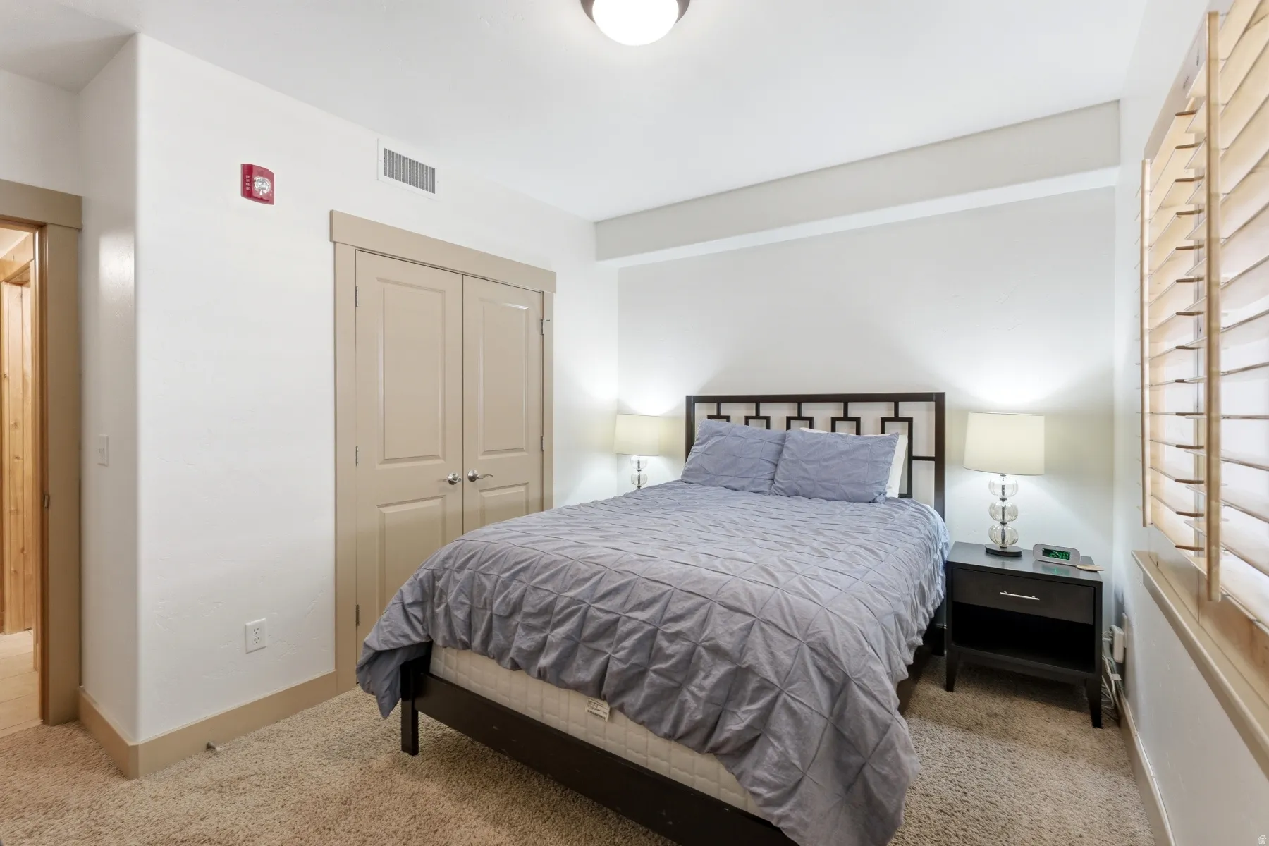 Carpeted bedroom with a built-in closet featuring double doors, light-colored plantation shutters, a contemporary ceiling light fixture, and neutral wall paint