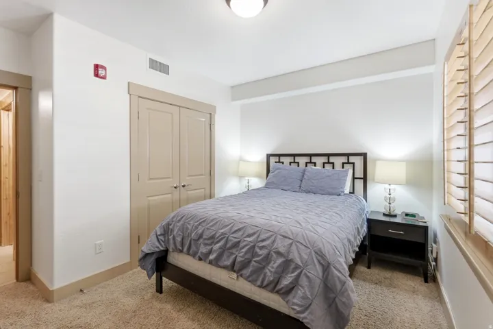 Carpeted bedroom with a built-in closet featuring double doors, light-colored plantation shutters, a contemporary ceiling light fixture, and neutral wall paint