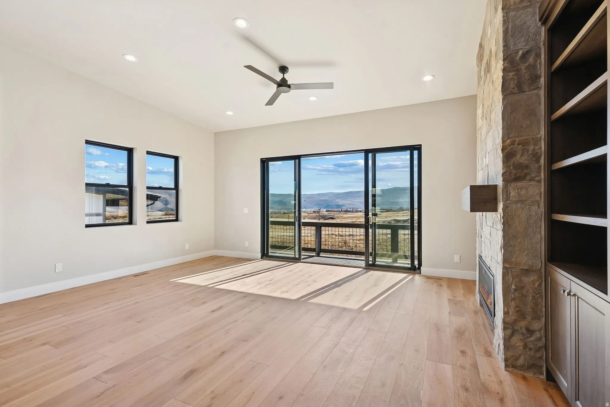 Bright interior featuring a stone-clad fireplace with built-in shelving, wood-finish flooring, and a ceiling fan