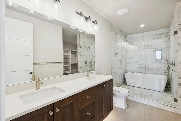 Bathroom featuring a dual vanity with white countertops and dark wood cabinetry, a frameless glass shower enclosure, a freestanding soaking tub, and marble-look tile walls