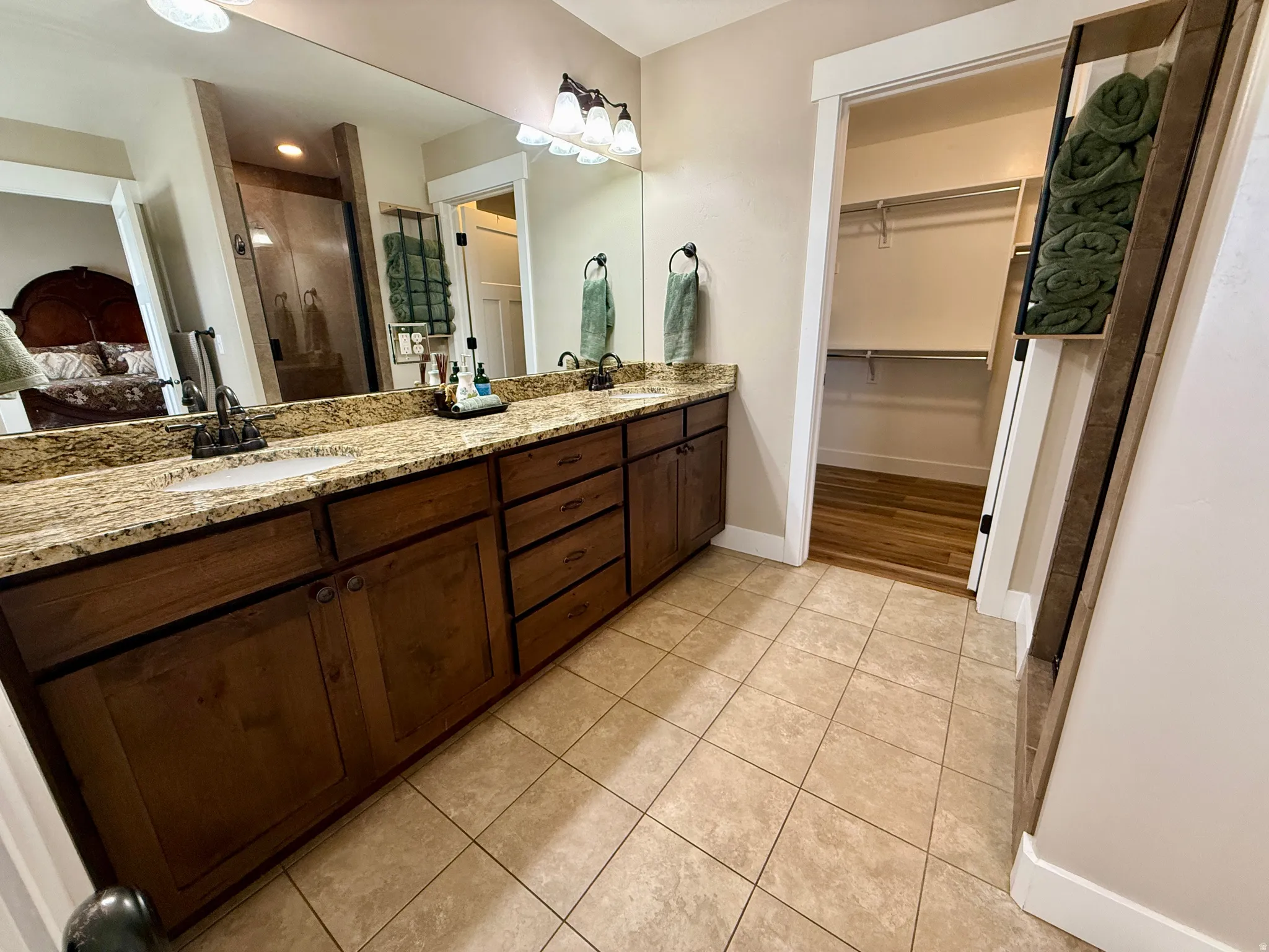Bathroom featuring a dual vanity with granite countertop and undermount sinks, wood cabinetry, tile flooring, and a walk-in closet with wood-finish flooring