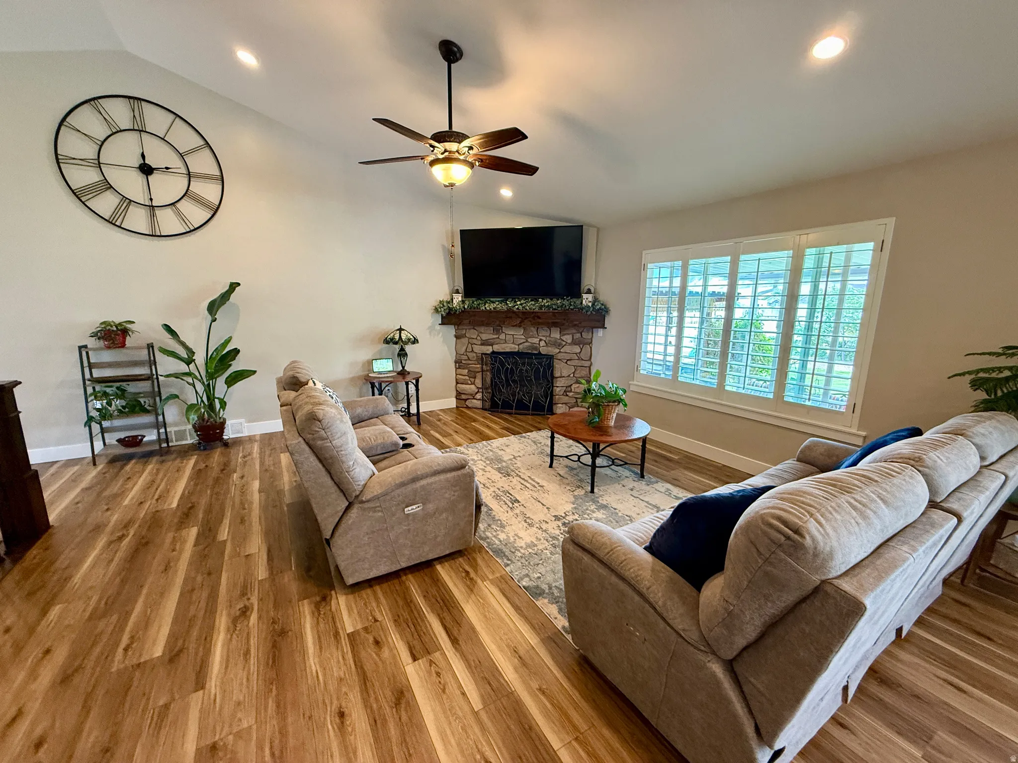 Spacious living area featuring vaulted ceilings with recessed lighting, wood-finish flooring, a stone-faced fireplace with a mantel, a ceiling fan with light fixture, and plantation shutters on the window