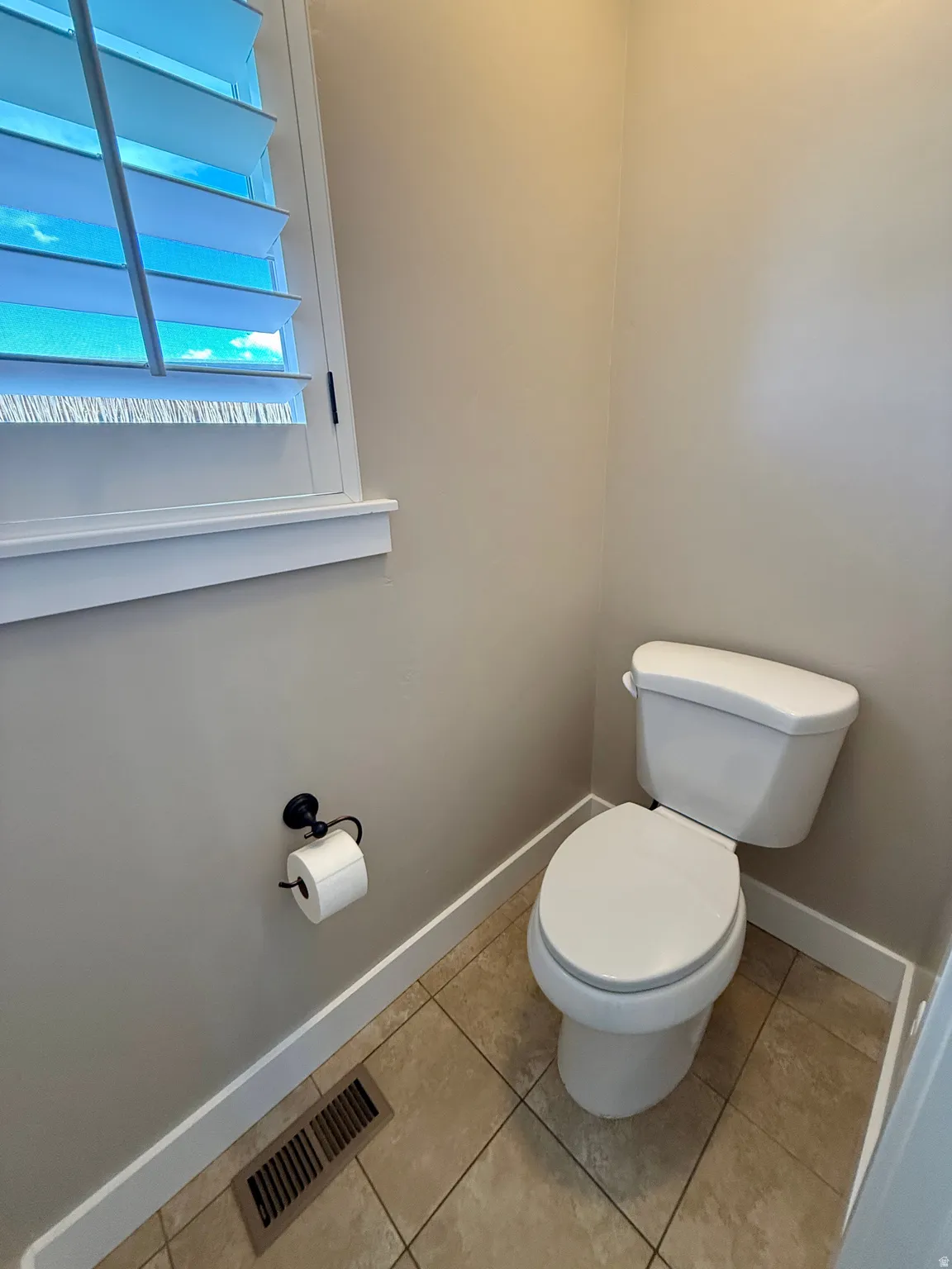 Powder room featuring a window with white louvered shutters, neutral-toned walls, tiled flooring, and a white two-piece toilet