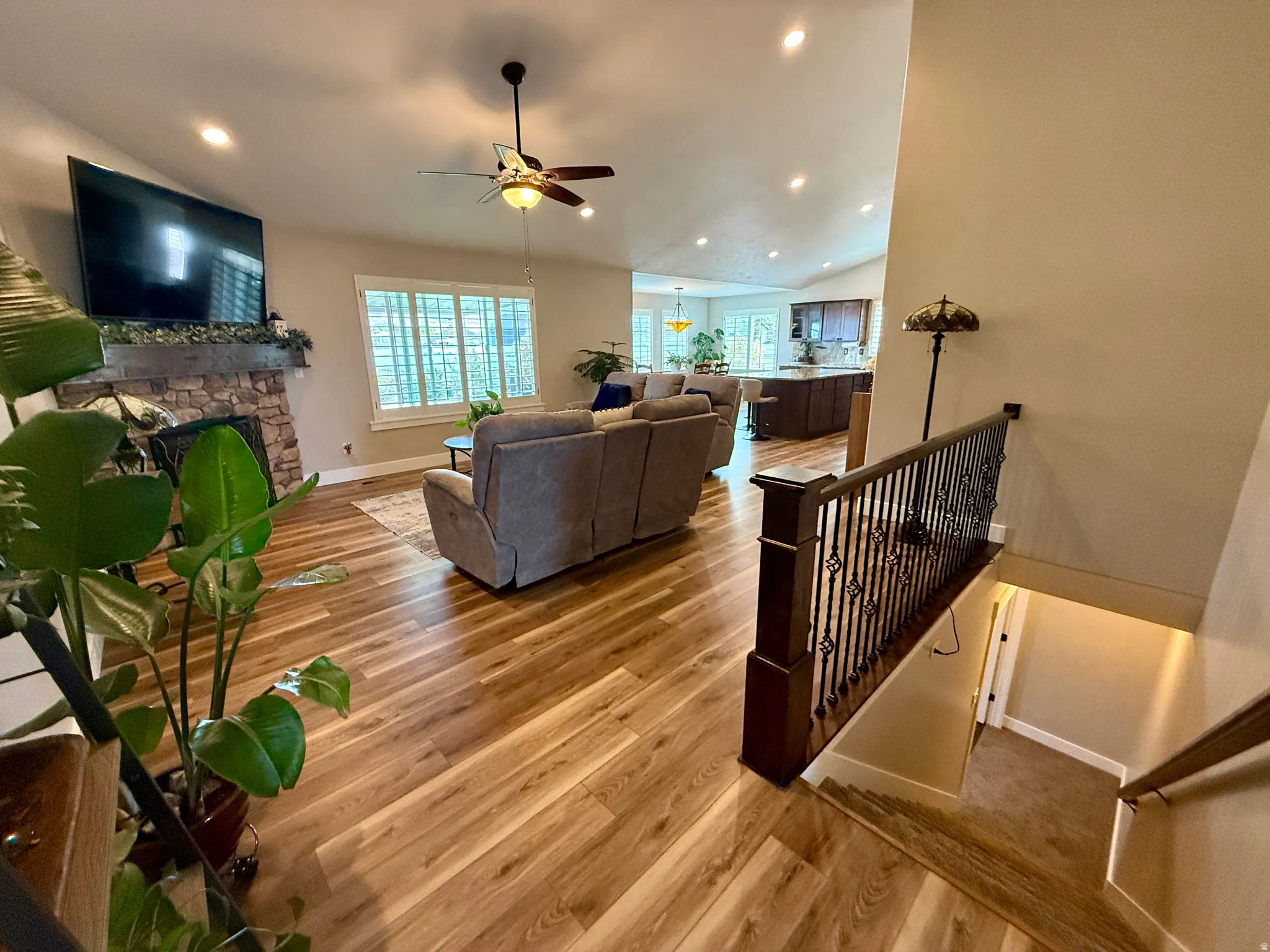 Bright open-concept living area featuring wood-finish flooring, a stone-faced fireplace, recessed lighting, and a ceiling fan