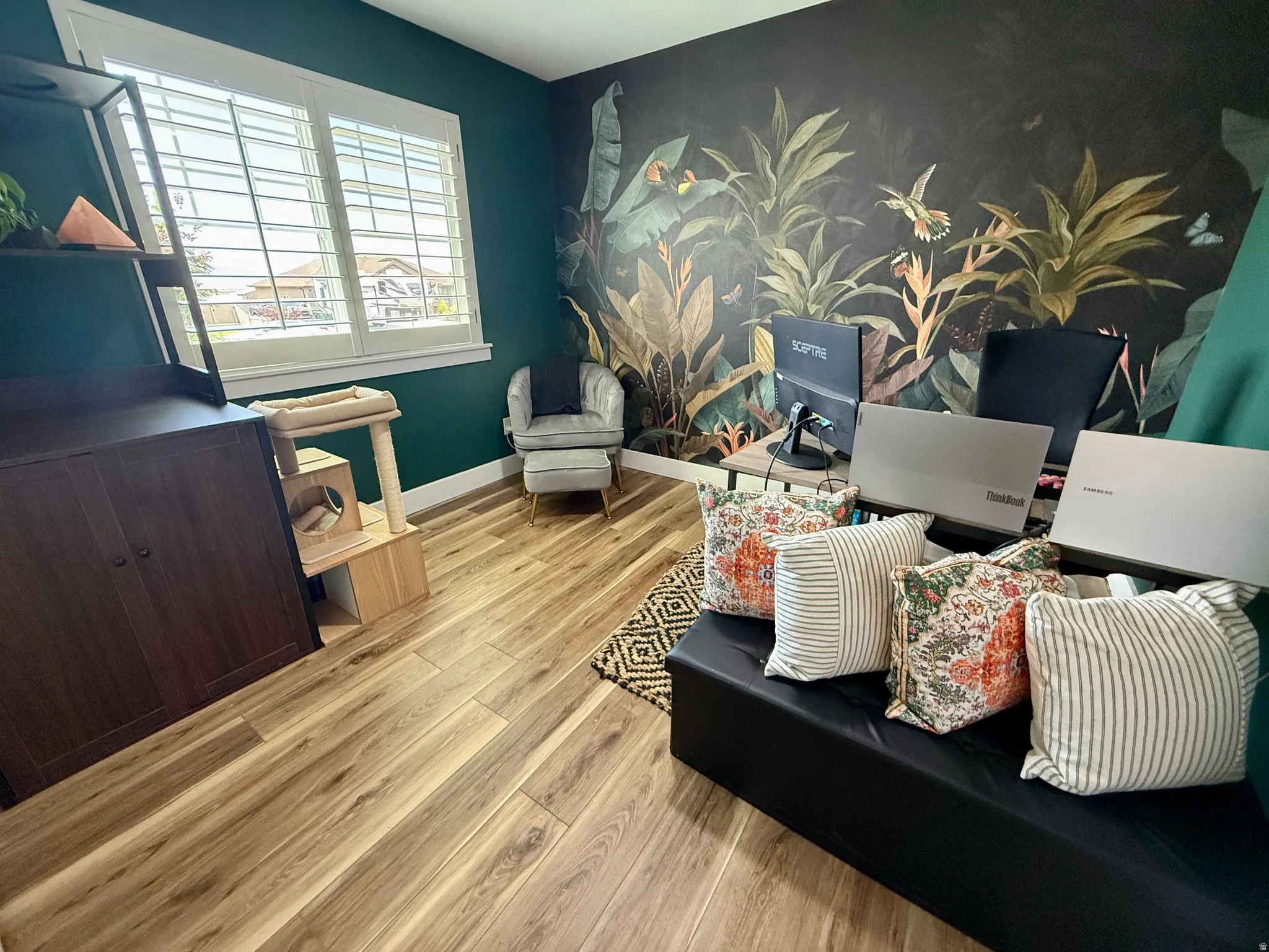 Interior room featuring wood-finish flooring, a large window with white plantation shutters, and a dark green accent wall with botanical wallpaper