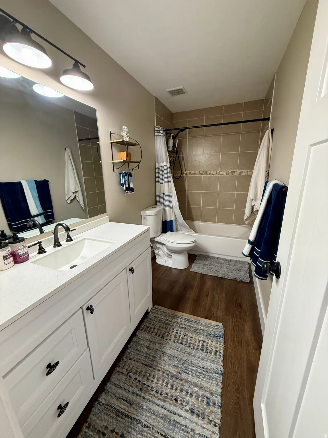 Bathroom featuring a white vanity with an integrated sink and dark hardware, wood-finish flooring, and a tiled shower with a decorative accent band
