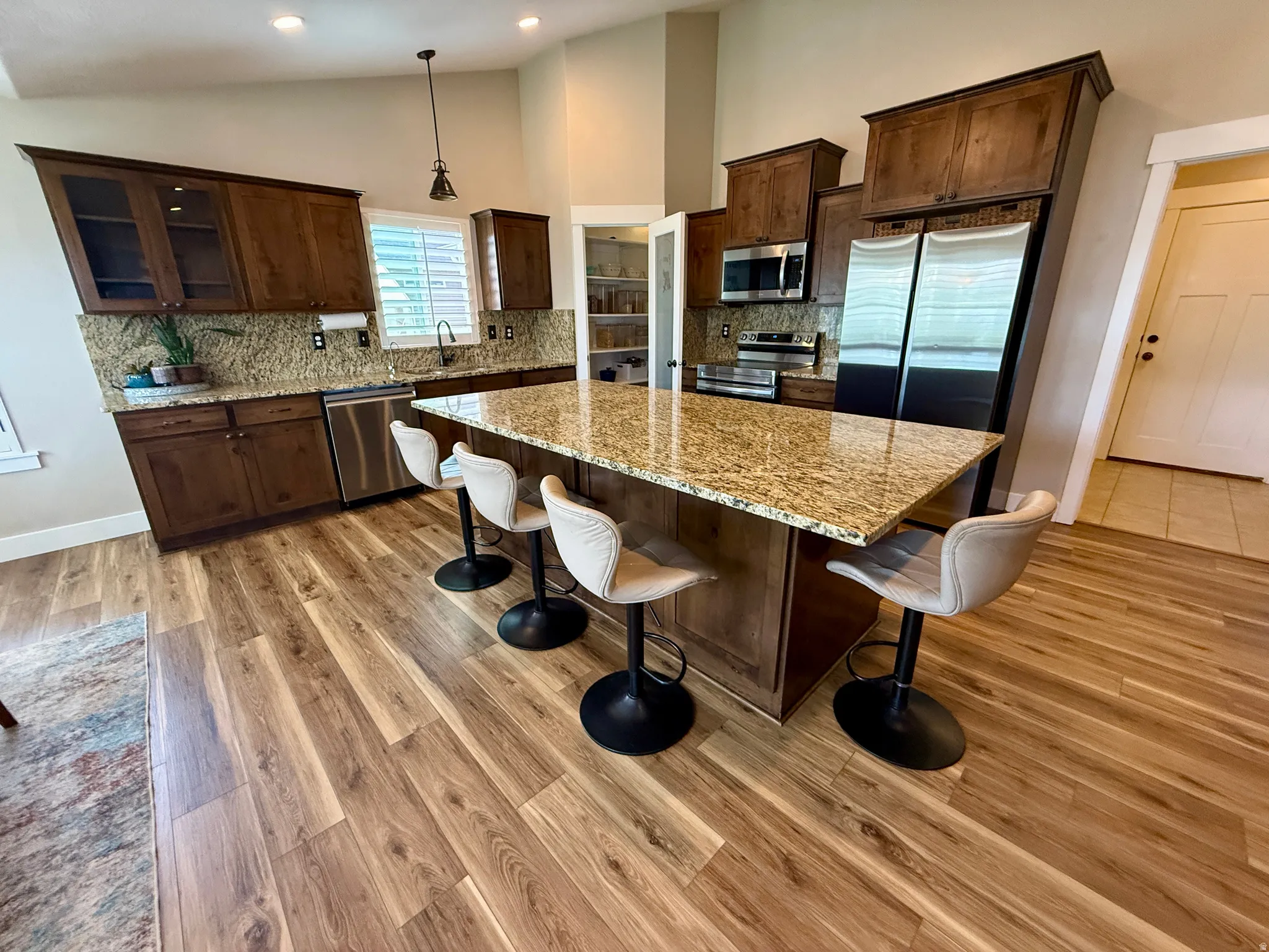 Expansive kitchen featuring a large island with a stone countertop, wood-finish flooring, dark wood cabinetry, stainless steel appliances, and a built-in pantry