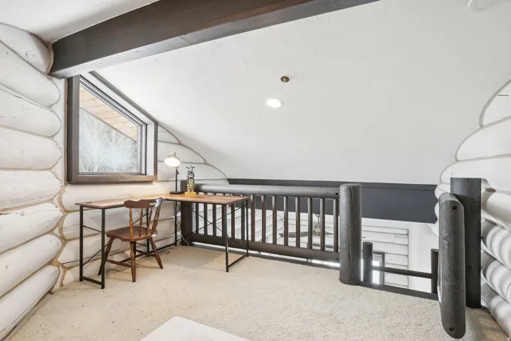 Loft area featuring white log walls, a dark wood-framed window, and exposed wood ceiling beams