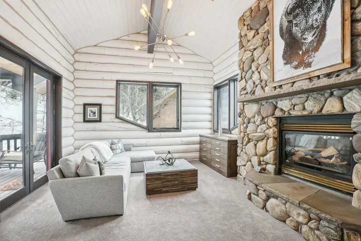 Vaulted ceiling with exposed wood beams, a stone-clad fireplace with a hearth, full-light French doors, and rustic wood-finish walls