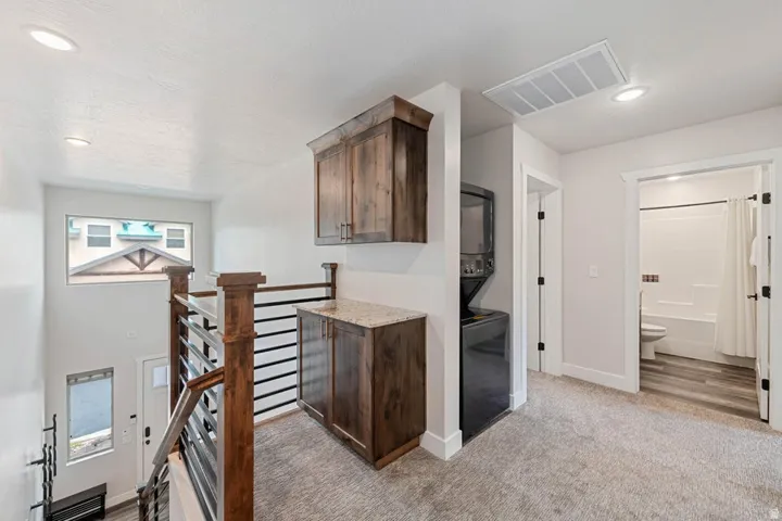 Upper-level landing featuring wood-finish cabinetry with a light-toned countertop, a stacked laundry unit, and a full bathroom entry