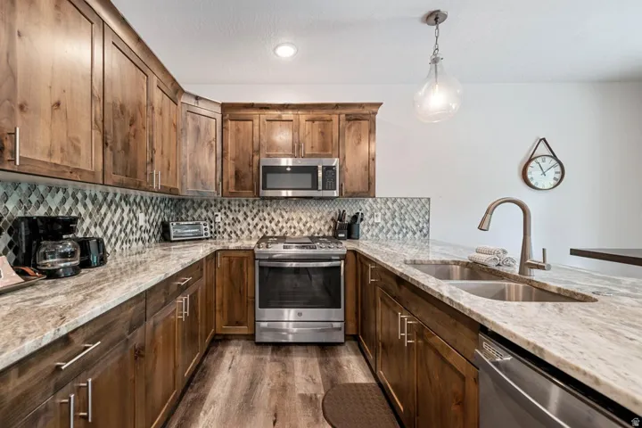Kitchen featuring wood cabinetry, light-toned stone countertops, a mosaic tile backsplash, stainless steel appliances, and wood-finish flooring