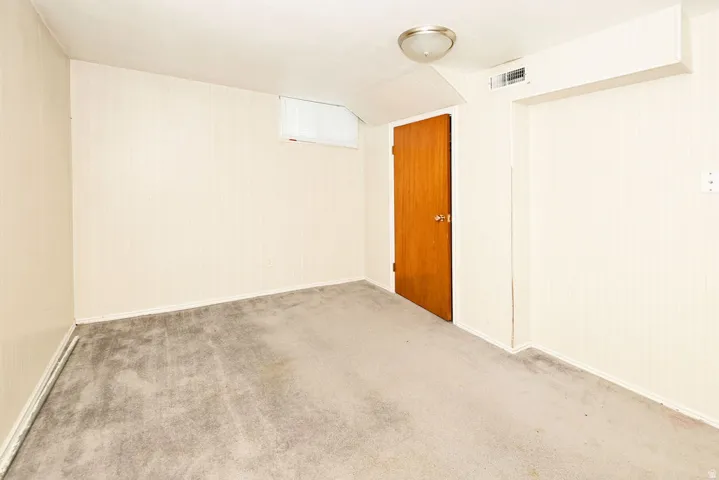 Carpeted room featuring a ceiling-mounted light fixture, a light wood-finish door, and a small, high-set window