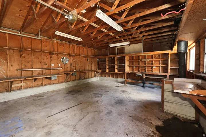 Spacious garage featuring exposed wood beam ceiling, concrete flooring, extensive built-in shelving, multiple work surfaces, and overhead fluorescent lighting