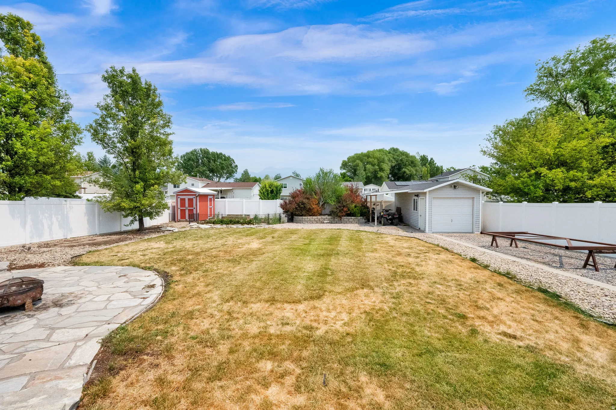 Expansive backyard featuring a stone patio, mature trees, a red storage shed, a detached garage, and white perimeter fencing