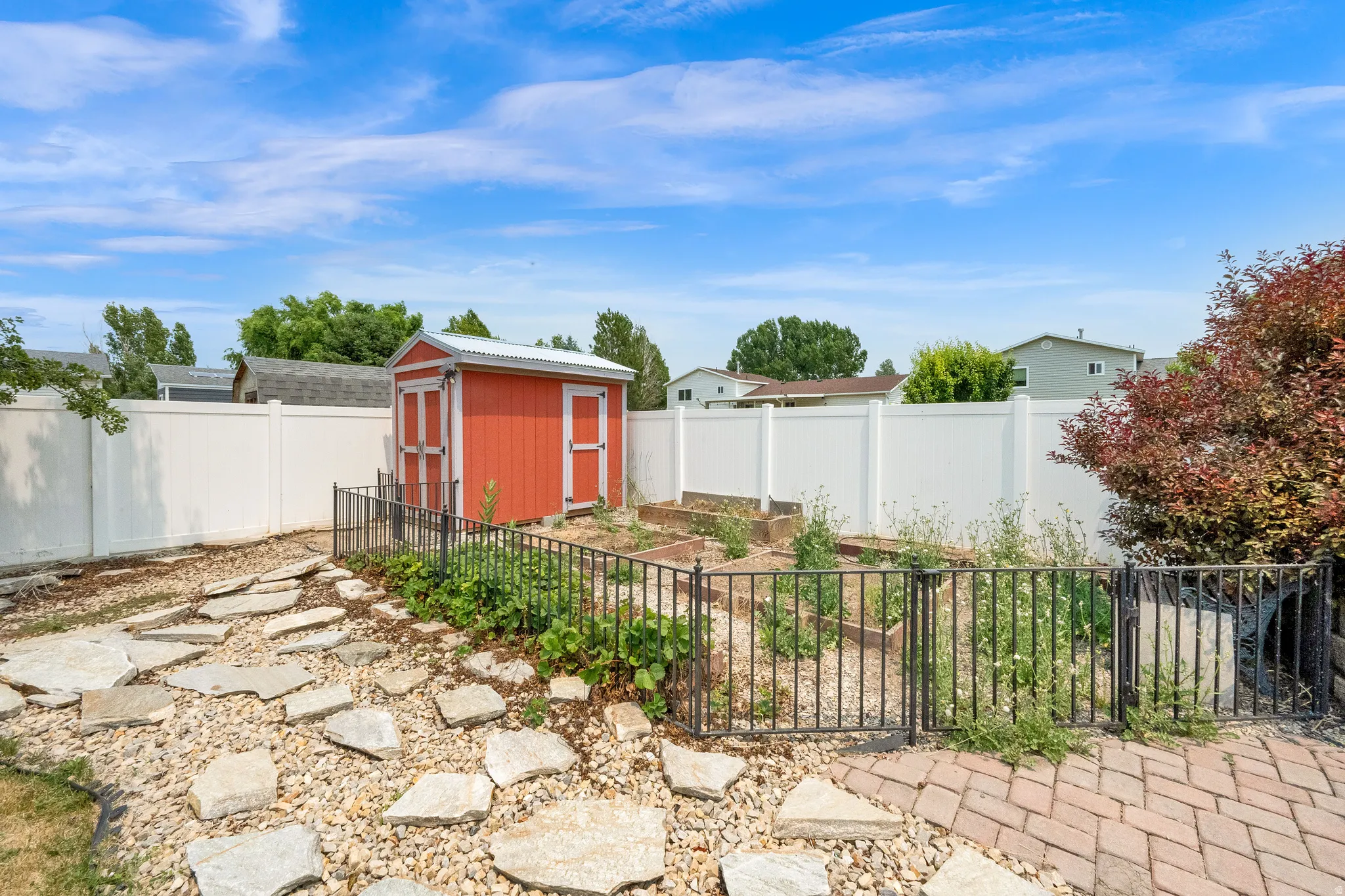 Backyard featuring a red storage shed, raised garden beds enclosed by a metal fence, a white privacy fence, a stone pathway, and brick patio elements