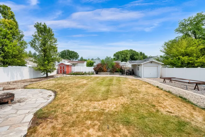 Expansive backyard featuring a stone patio, mature trees, a red storage shed, a detached garage, and white perimeter fencing